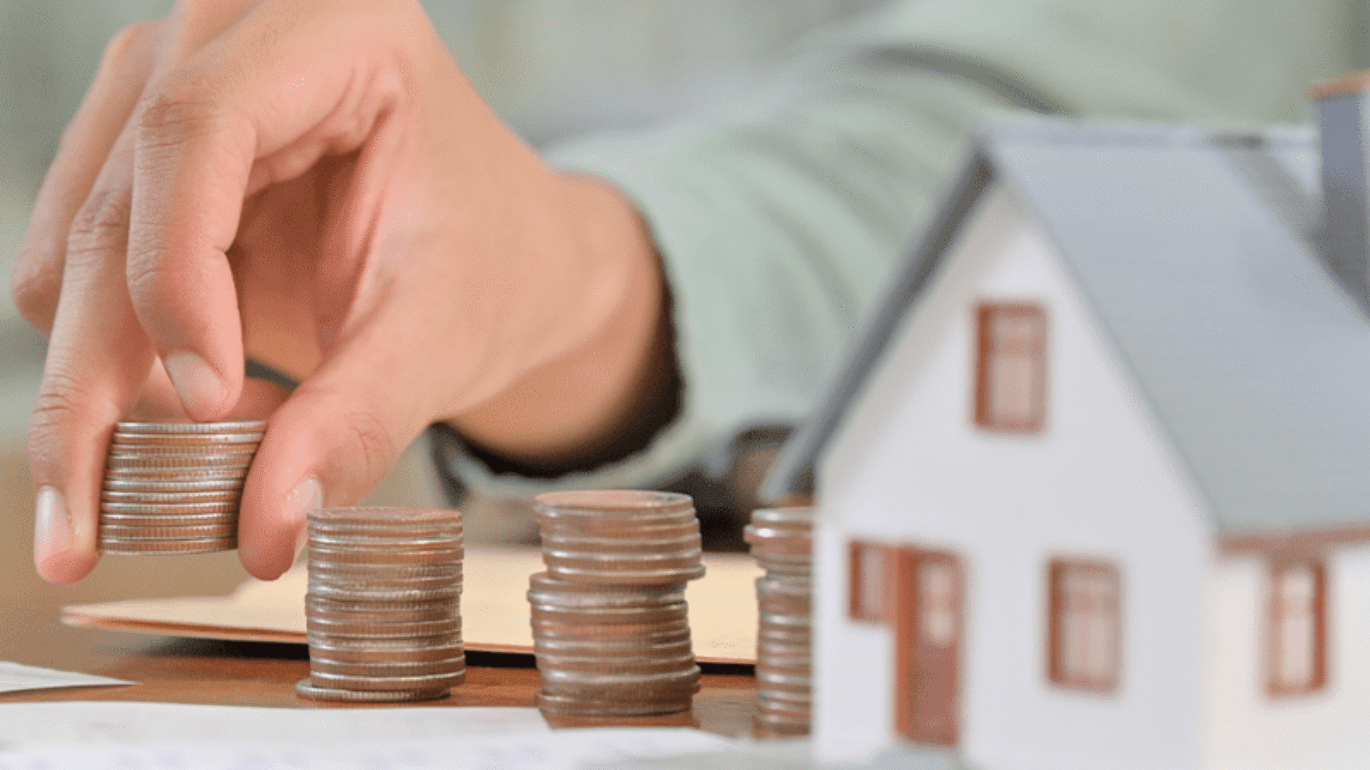 Hand stacking coins beside a model house representing home building costs