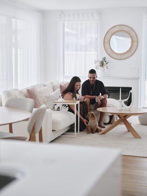 A woman pats a scruffy dog while sitting with a man on a white couch in the living room
