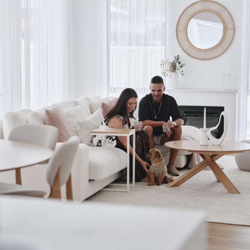 A woman pats a scruffy dog while sitting with a man on a white couch in the living room