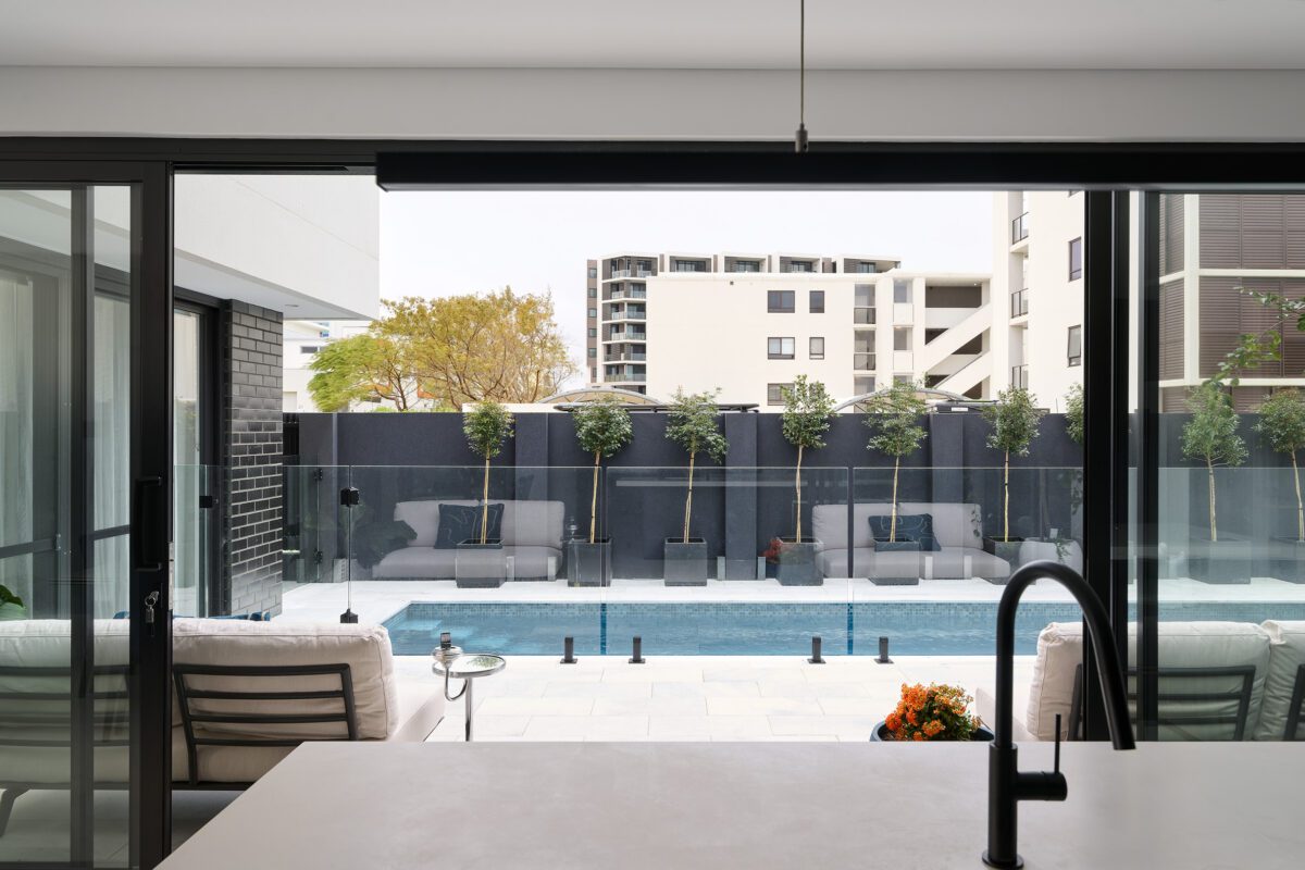 The photo shows the view from the kitchen island out toward an outdoor entertaining area with a glistening blue swimming pool for our North Coogee clients. Our clients share their experience and review Residential Attitudes.